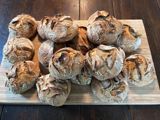 Auf einem dunklen Holztisch liegt ein Holzbrett. Darauf liegen zahlreiche frisch gebackene Brötchen mit rustikalem Ausbund