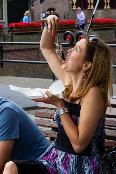 A young woman is sitting on a bench in The Hague. She is eating a herring by holding it up by the tail with her right hand and then eating off pieces with her mouth. But in this photo the fish is still hanging above her head, her mouth open. There are small pieces of raw onion on the fish. She is photographed from the side. From just below her waist. Her right little finger is elegantly raised. In her left hand she holds a white bowl. In the background in the distance the railing of the "Vijverhof" in The Hague.  A few people are walking by. She has bare arms, it is summer and nice weather.