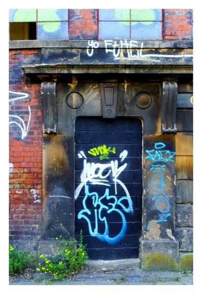 An old, bricked-up doorway sits in the center of a weathered, soot-stained stone facade on a red brick wall. The doorway is heavily marked with vibrant graffiti in blue, white, and neon yellow. Above it, blackened stone pillars frame the doorway with decorative round motifs and a keystone detail. Faded graffiti and grime cover the wall, while cracked glass windows with green blotches sit above. Wild yellow flowers and weeds grow from cracks at the base, hinting at abandonment. The image has a soft focus and lifted shadows, giving it a dreamy, film-like quality with a sense of urban decay.
