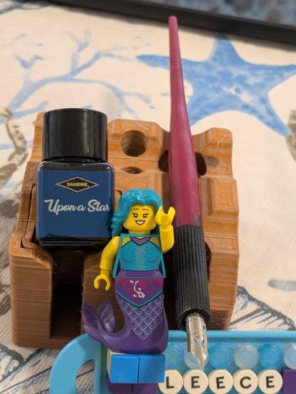 A little Lego mermaid sits on a badge constructed of Lego, with 'LEECE' formed on it. To the rear an inkstand with a bottle of " Upon A Star" ink and a dip pen.