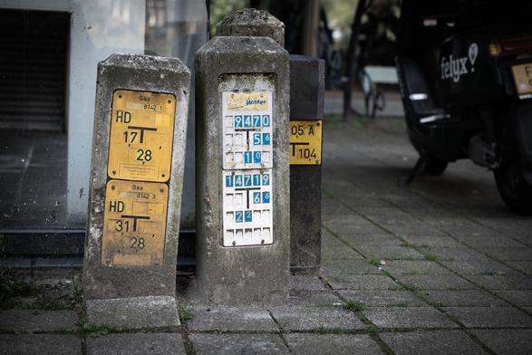 Utility location sings on beton posts, slightly tilted, positioned around a building corner of glass and steel. [Nikkor Z 24–120mm f/4.0 S auf Nikon Z5]