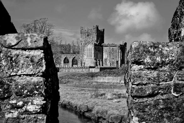 A black and white photograph of Tintern Abbey in County Wexford, Ireland, framed between two stone walls in the foreground. The ancient abbey, with its tall, arched windows and crumbling facade, stands majestically on a grassy rise. A narrow river flows in the immediate foreground, reflecting the light. The sky above is partially cloudy, and a few bare trees are visible in the background to the left of the abbey. The overall mood is historic, serene, and evocative.