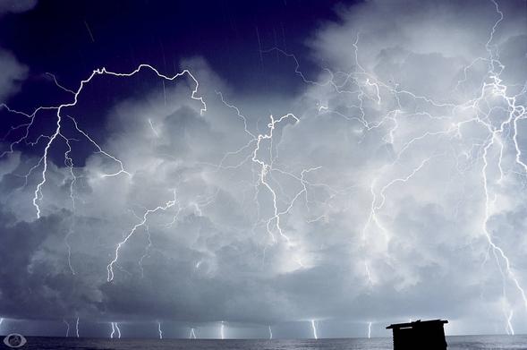 The sea is only a thin strip with a black square at the lower right which is a cabin and then thousands of lighting bolts in and out of the clouds. where the clouds clear (upper left) star trails are visible.