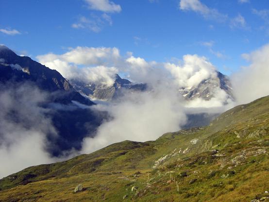 Wolkenverhangener Blick aus dem Maderaner Tal in das Gotthardtal, im Vordergrund sind grüne Almweiden zu sehen.