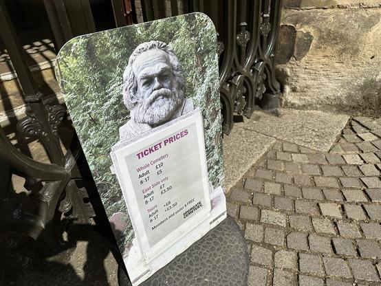 Photo of a board with times and prices. The background is a photo of Karl Marx’s gravestone