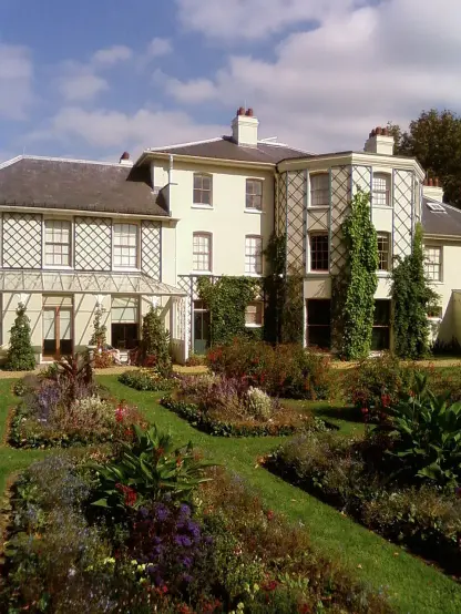 The image depicts a large, multi-story house with a garden in front. The house features a combination of architectural styles, including traditional elements like a gabled roof and some sections with decorative timber framing. The garden has well-maintained flower beds and lush greenery, contributing to a picturesque setting. The sky appears to be partly cloudy, suggesting a pleasant day.

Image Credits: Wikimedia / Anthonyeatworld at English Wikipedia / CC BY-SA 3.0