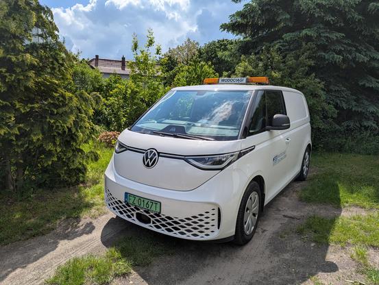 Biały elektryczny van VW ID.Buzz widziany z przodu. Na dachu kogut wodociągów. Tło - zielone krzaki.