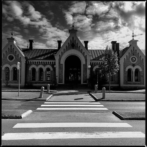Square monochrome photo of a stone train station house taken across a cross-walk making the image symetrical, the cloudy but blue sky can be seen above it, the photo has a Hasselblad frame