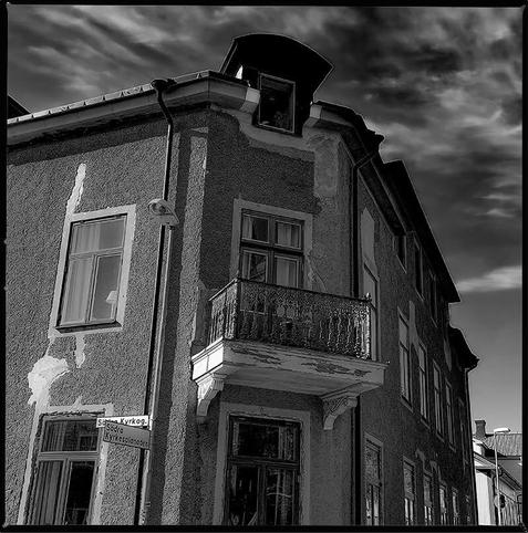 Square monochrome photo of the corner of a stone house with a balcony and windows, the cloudy but blue sky can be seen above the roof and a little to the right side, the photo has a Hasselblad frame