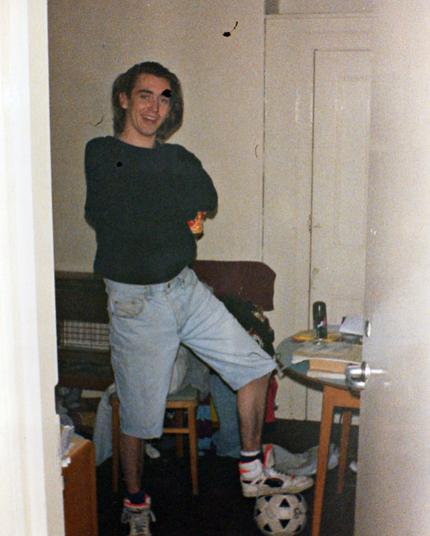 Scan of a colour photograph in portrait orientation. We're looking through the doorway into the bedroom of a young man. The lighting in the room suggest the curtains are drawn and the light is on so it's probably early evening. The man is posing with one foot resting on a football on the floor. He's wearing trainers, has light denim jeans cut off just at the knee, and is wearing a black jumper. His arms are cross and he's smiling at the camera, and there's a can of drink in one hand just visible under his armpit. The room looks quite messy. There's a gas fire on the wall behind the man and there's part of a circular table visible to the right of him, partly hidden by the open door. On the table there is some deodorant, some books, and papers on the table and on a chair behind it - and on the floor - there is discarded clothing.
