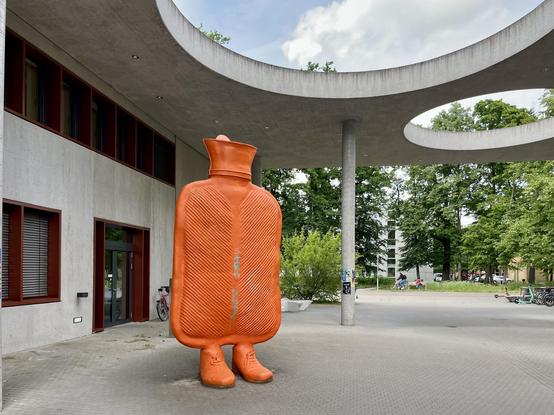 The image features a large, orange, hot water bottle sculpture with human-like legs, standing in an outdoor plaza. The sculpture is textured with ridges, resembling the surface of a real hot water bottle. It is positioned in front of a modern building with a concrete structure and circular openings in the ceiling, providing shade. The building has a glass entrance and windows with red frames. In the background, there are trees and a bicycle parked nearby. The plaza is paved with grey tiles, and there are people and bicycles visible in the distance. The sky is partly cloudy, suggesting a pleasant day.

Provided by @altbot, generated privately and locally using Ovis2-8B

🌱 Energy used: 0.147 Wh