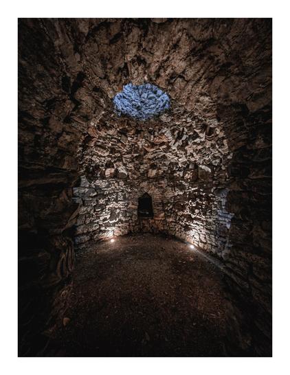 The photo shows the interior of the ghiacciaia. It's a circular, stone-walled chamber. The walls are made of rough, uneven stones, and there's a small opening in the ceiling letting in a bit of light.  The floor is made of packed earth.  Two small lights are placed on the floor near the wall, illuminating part of the space.