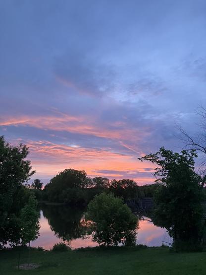 Dark blue grey clouds ombré down into orange and pink predawn sky over a treeline and reflected in the river in the foreground