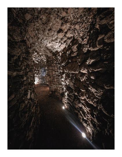 The photo is a dimly lit, narrow chamber within a stone structure. The walls are made of rough, uneven stones - just like the bigger chamber. It looks like a narrow passageway but is actually a chamber and leads nowhere. There are recessed lights in the ground that light the way.