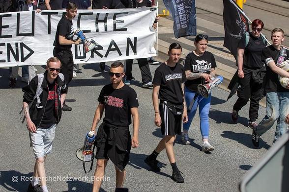 Vor einem schwarz-weißen Transparent „Deutsche Jugend Voran“ steht Justin Elbel mit Megafon und einem schwarzen Shirt im Stil von New Balance mit der  Aufschrift „NB nazi bande“. Davor laufen Stella Schmidt, mit Megafon und einem Lonsdale-Shirt, Anthony Scholz mit einem Shirt „Anti-Antifa“ und Julian Milz mit Mgefaon und einem Shirt „Division Berlin“.