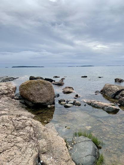 A rocky shoreline by a calm sea under a cloudy sky. Large boulders and smaller stones are visible along the water's edge, with distant islands in the background.