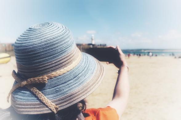 A picture of a woman wearing a hat taken from behind. It's a close up, only her shoulder and hat are visible. She is facing away from camera holding up a phone, taking a photo of a beach with the sea in the distance.