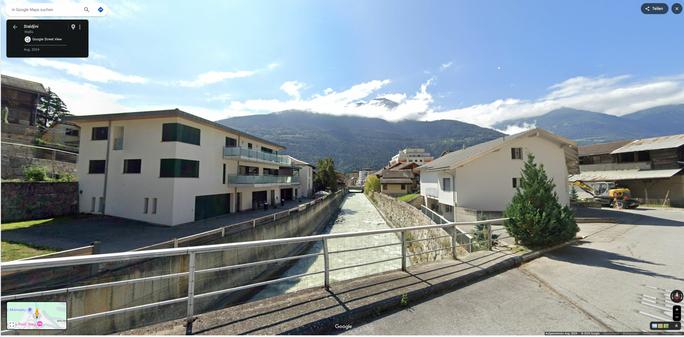 Ein Google streetview Screenshot aus der Gemeinde Gampel Steg. Aufgenommen auf der ersten Brücke im Gemeindegebiet über die Lonza, flußabwärts. Links und rechts des gemauerten Gerinnes Häuser.