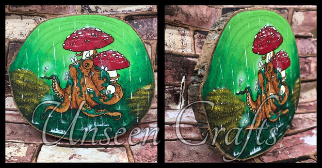 Two photos, one showing side view, one front view. Oval wood slice with bark edge and burned design showing an orange octopus with green mossy bits and tiny white mushrooms growing on her. She sits under two red and white amanita mushrooms. There are plants in tones of brown and yellow that resemble gorse at either side. It is raining, the rain falls down across the scene and splashes on the deep green floor underneath the octopus. The huger background is a more vibrant green.