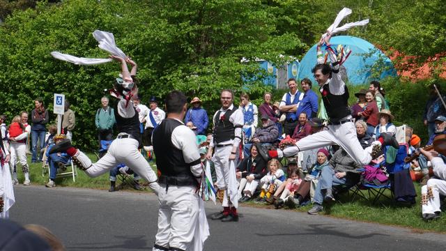 Three morris dancers with white shirts and trousers and black vests, decorated with multicolor ribbons, dancing on a paved surface. Three and standing and two are in motion, leaping high with arms raised. Behind them is an audience including a number of other morris dancers.