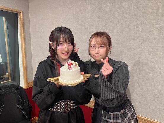 Two women are posing indoors with a celebratory cake. The woman on the left, wearing a black hoodie with braided hair and round glasses, is holding the cake decorated with strawberries and flowers. The woman on the right, dressed in a gray top and plaid skirt, is making a heart gesture with her fingers and smiling at the camera.