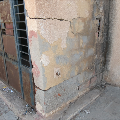Foto vom Eingangsbereich der Karasor-Schule, außen. Die Eingangstür besteht aus verrostetem Metall. Die Mauer hat Risse und schwarze Verfärbungen an der Fassade.