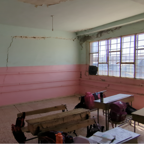 Foto von einem Klassenraum. Farbe ist abgebröckelt. Die obere Hälfte der Wand und die Decke sind in einem hellen grün gestrichen. die untere Hälfte der Wand ist rosa. Die Wände weisen Risse auf. Die Schulbänke und Tische sind aus Holz.
