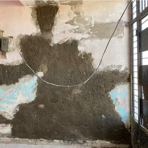 Foto vom Eingangsbereich der Karasor-Schule, innen. Der Putz ist abgebröckelt. Ein Kaputter Elektronik-Kasten hängt an der Wand und ein isoliertes Kabel hängt an der Wand und führt zum oberen Bereich der Eingangstür. Mutmaßlich ist das die Stromversorgung für die Eingangsbeleuchtung.