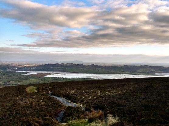 The view is across a dark heath landscape from Knocknarea in the foreground with a narrow path into the distance across the Sligo countryside with bogs, bays and a range of hills in the distance under an atmospheric sky.