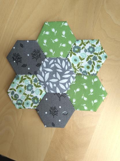 Seven hexagons sewed together. The middle one is grey with white leaves, two are dark grey with little trees, two are green with little white flowers, two are white with green and teal flowers