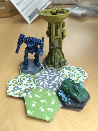 Same piece but with a blue half-painted battlemech, a green tank, and a radio tower on hex bases placed on individual fields like it was a game map