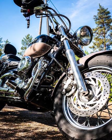 Chrome and black motorcycle parked outdoors with disc brake on the front wheel, shiny chrome engine parts, and handlebars with leather grips. Sunlight reflects off metal surfaces under a clear blue sky, with tall trees in the background.
