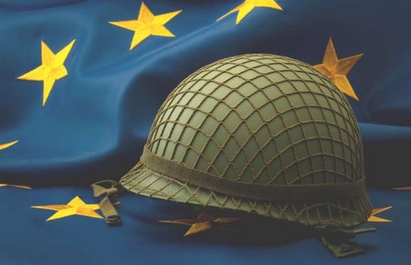 L'immagine mostra un elmetto militare americano della seconda guerra mondiale appoggiato su una bandiera europea. L'elmetto è di colore verde oliva e presenta una rete mimetica. La bandiera è blu con dodici stelle dorate a cinque punte disposte in cerchio.

Descrizione generata con AI.