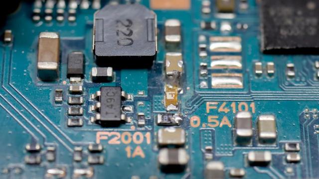 Makroaufnahme einer Platine mit winzigen SMD-Bauteilen. In der Mitte ist eine SMD-Sicherung, die vor dem Einbau noch deutlich "gesünder" aussah. Auch die Lötstellen von benachbarten SMD-Bauteilen sehen beschädigt aus.