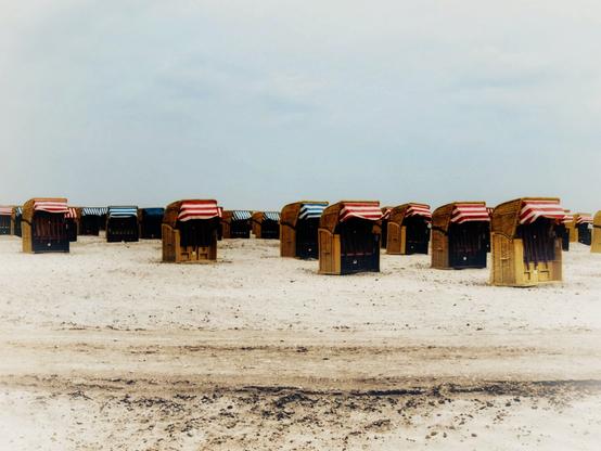 A wide, slightly desaturated shot shows numerous hooded beach chairs, known as "Strandkörbe", arranged in rows across a sandy expanse under a pale, overcast sky. The chairs are predominantly made of woven wicker with dark interiors and canopies featuring striped fabric in various colour combinations, most prominently red and white, but also blue and white, and black and white. The sand in the foreground shows some darker, damp patches. The horizon line where the sand meets the sky is low in the frame, emphasizing the vastness of the beach and the number of the beach chairs. The overall atmosphere is calm and slightly muted.