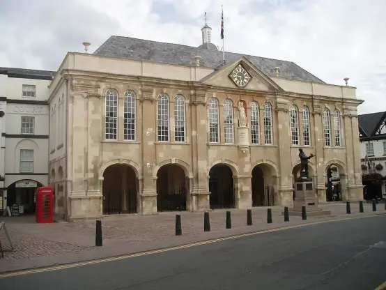 This image appears to depict a historic building, likely a town hall or similar civic structure. It features a classical architectural style with large windows, columns, and a prominent clock. There are also elements such as a statue in front of the building and traditional elements like a telephone booth nearby, indicating a blend of historical and modern features. The street appears to be paved, suggesting a pedestrian-friendly area.

Image Credits: Wikimedia / ShireHallMonmouth / CC BY-SA 3.0