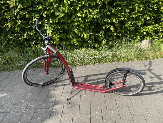 Photo of a red footbike (kickbike/scooter) for adults with 26“ front wheel and 20“ rear wheel. In the back there is an hedgerow.