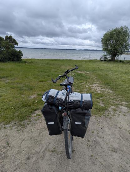Rower z sakwami stojący na zielonym terenie nad brzegiem jeziora. Wyburzona tafla wody i chmury na niebie.