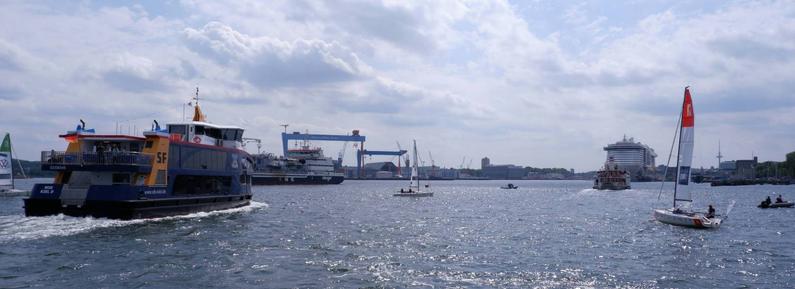 Blick aufs Wasser mit vielen Schiffen, Segelbooten und paddlern. Im Hintergrund sind Werftkräne zu sehen.