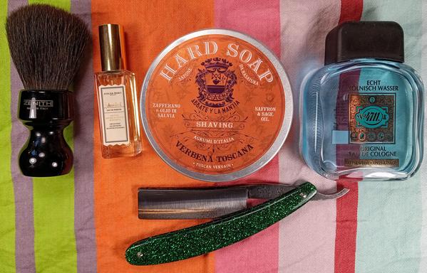 (Le Chat Mistral description)

The image showcases a collection of grooming items arranged on a striped background. On the left, there's a black shaving brush with a black handle. Next to it is a small bottle of "Atelier Reboul" perfume with a gold cap. In the centre, there's a round tub of "Verbena Toscana" shaving soap with an ornate label. To the right of the soap, there's a bottle of "4711 Echt Kölnisch Wasser" cologne with a blue liquid inside. At the bottom, there's a straight razor with a green glittery handle. The items are neatly laid out, suggesting a traditional shaving setup.