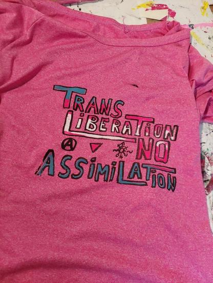 A pink t-shirt. A print on it reads 'Trans Liberation, No Assimilation.' The font is hand-written, and has a black outline, and a trans-flag fill-out over the whole text.