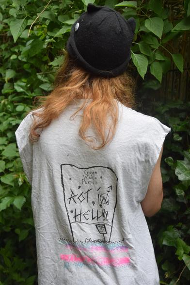 Photo of a person with long hard from the back, wearing a light grey shirt with cropped sleeves, that has a print on it showing a protest, printed in trans flag colours, which carries a big banner (printed in black) above their heads that reads: Green, White, Purple, Rot In Hell.