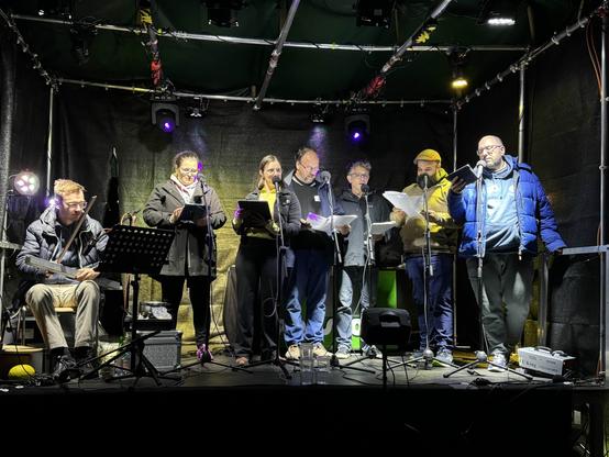 A small group of people, standing on stage, is engaged in a performance. They are holding scripts and are positioned near microphones. A musician, seated, plays an instrument. The stage is illuminated with colored lights, and the backdrop is dark fabric.