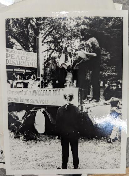 A black and white photograph.  A group of women are on a stage chanting with some trees in the background, and there are some banners and protest signs which read "Women's day of action for peace and disarmament" and "...in the event of a nuclear war there will be no survivors". A child looks on at the group of women - they are wearing a black bandanna around their head (or it is trick of lighting from a "mistake" ine photograph) which gives it the eerie quality of somebody's face, when it is actually just the back of their head.