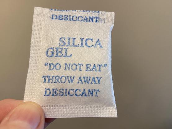 A thumb and forefinger holding a little white sachet labeled:
SILICA GEL
"DO NOT EAT"
THROW AWAY
DESICCANT