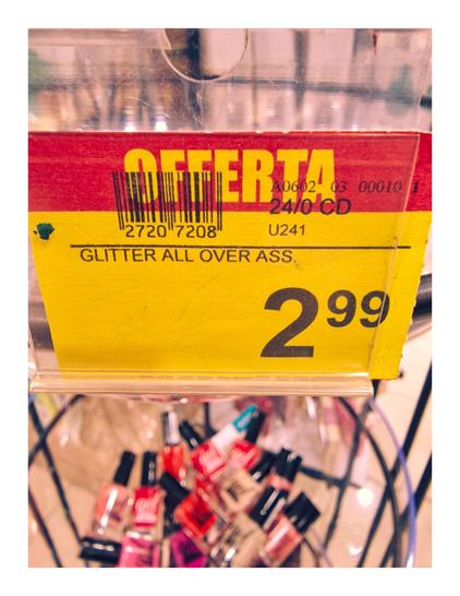 The photo is of a yellow and red shop display sign in Italian that reads "Offerta" (meaning "Offer" or "Sale") with a price of €2.99. Below the barcode, the product description reads "GLITTER ALL OVER ASS,". Which is in reference to the assorted nail polishes. The basket below the sign contains various bottles of these brightly colored nail polish.