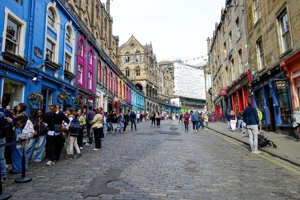 Cobblestone street bustling with pedestrians, lined with brightly colored buildings in shades of blue, pink, and red on the left. On the right, traditional stone buildings under a cloudy sky, marked by a historic brown stone structure with gothic architectural features at the end of the street. Banners decorate the street above, and a crowd queues in front of a cafe on the left.