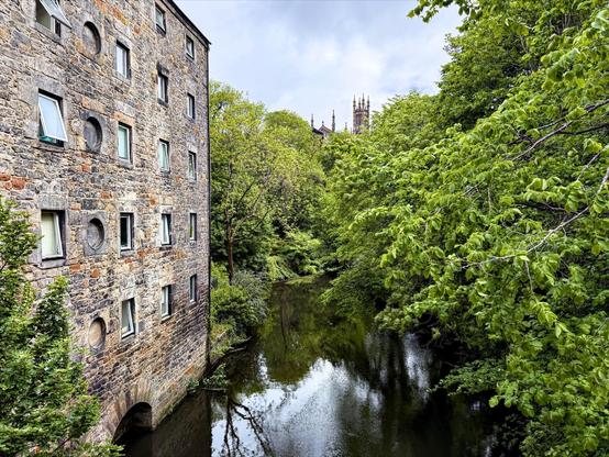 Old stone building with multiple windows overlooking a calm river, surrounded by lush green trees. Gothic spires visible above the treeline in the background.