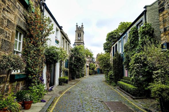 Cobblestone street lined with traditional stone houses, some covered in climbing plants, leads toward a church tower in the distance. Potted plants and small gardens enhance the quaint, greenery-filled scene.
