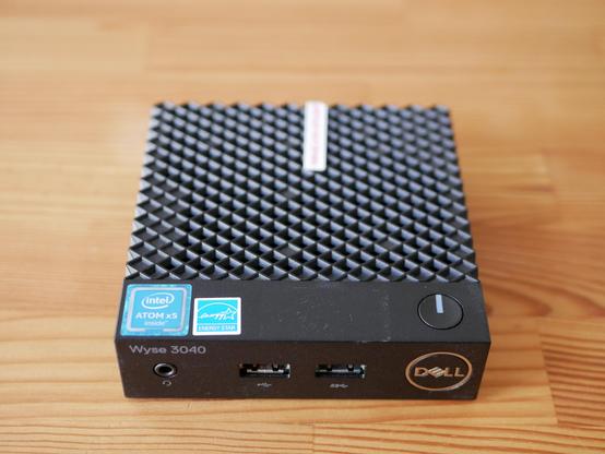 Foto eines rechteckigen Minicomputers. Das Gehäuse ist schwarz, oben hat er eine Gitteroberfläche, rechts oben vorne ist ein power button. Vorne links steht "Wyse 3040", rechts "DELL". Auf der Vorderseite sind drei ports: Audio und zwei USB ports. Er liegt auf einem Holztisch.